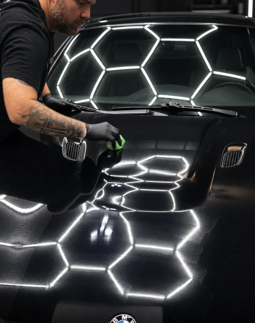 A TC Auto Solutions detailer applying ceramic coating to a black BMW bonnet under hexagonal bay lighting